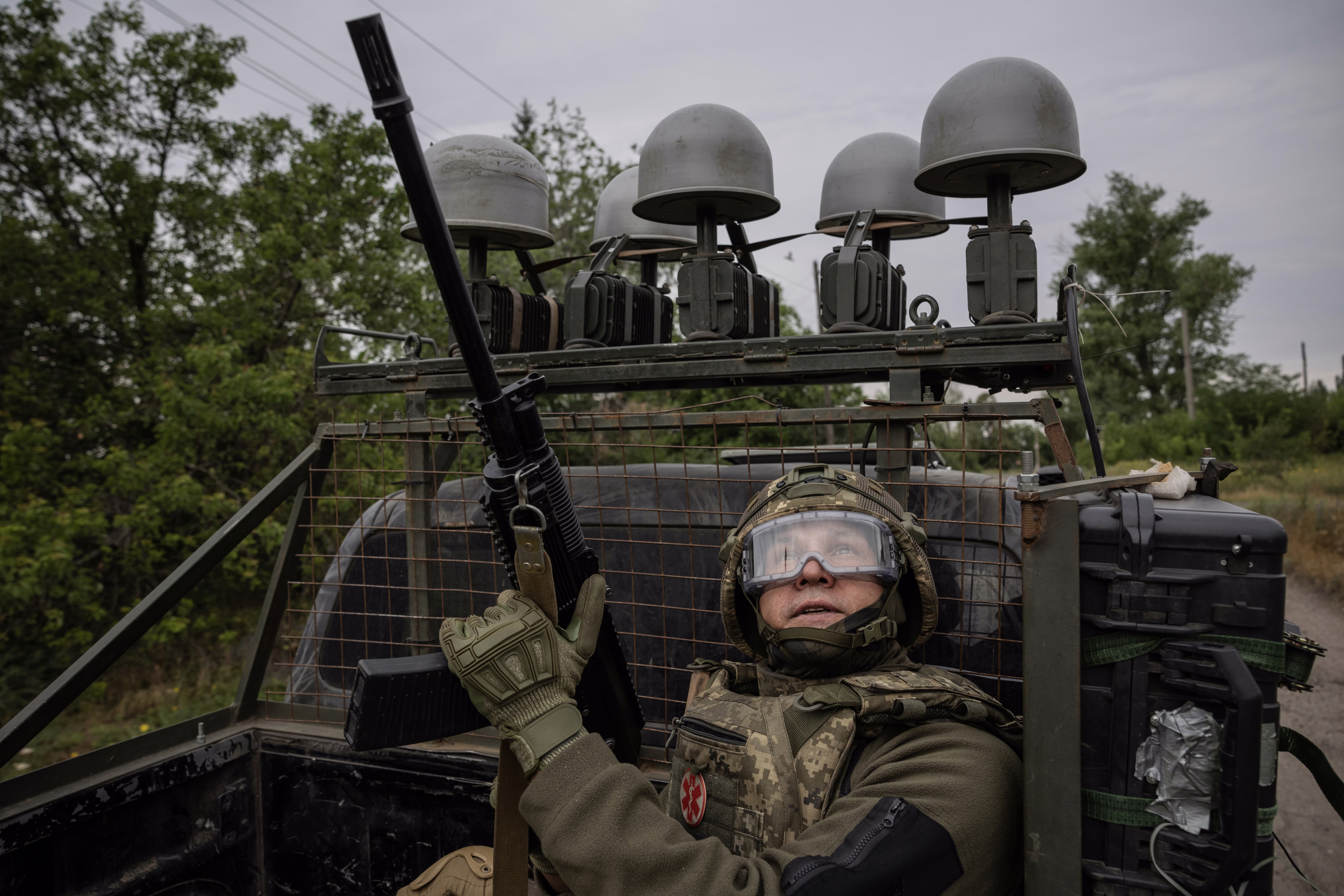 A soldier holds a weapon while in the bed of a military pickup truck, watching the sky for drones.