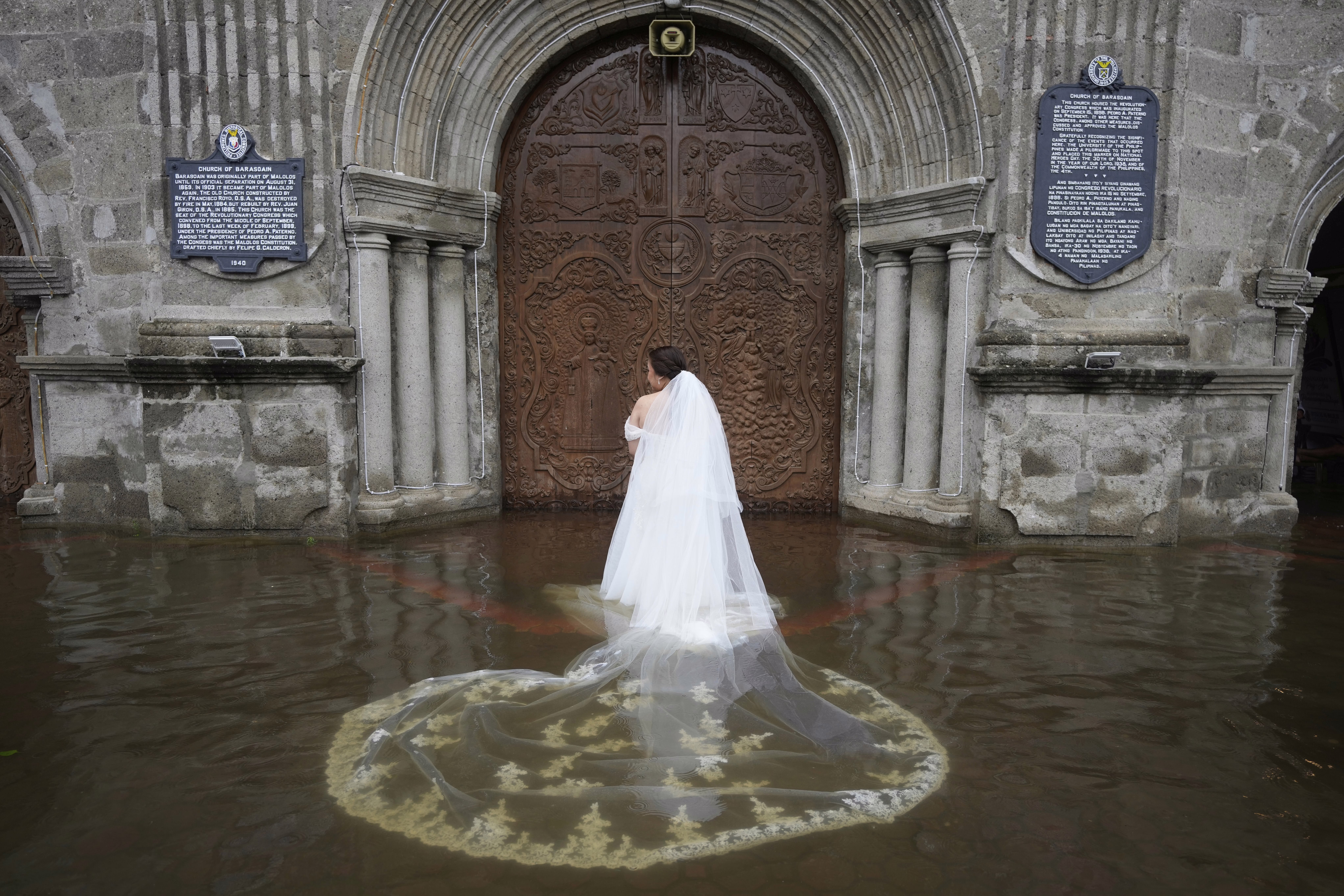 A bride poses for a photo outside a church, standing in ankle-deep floodwater, her long veil spread out in the water.