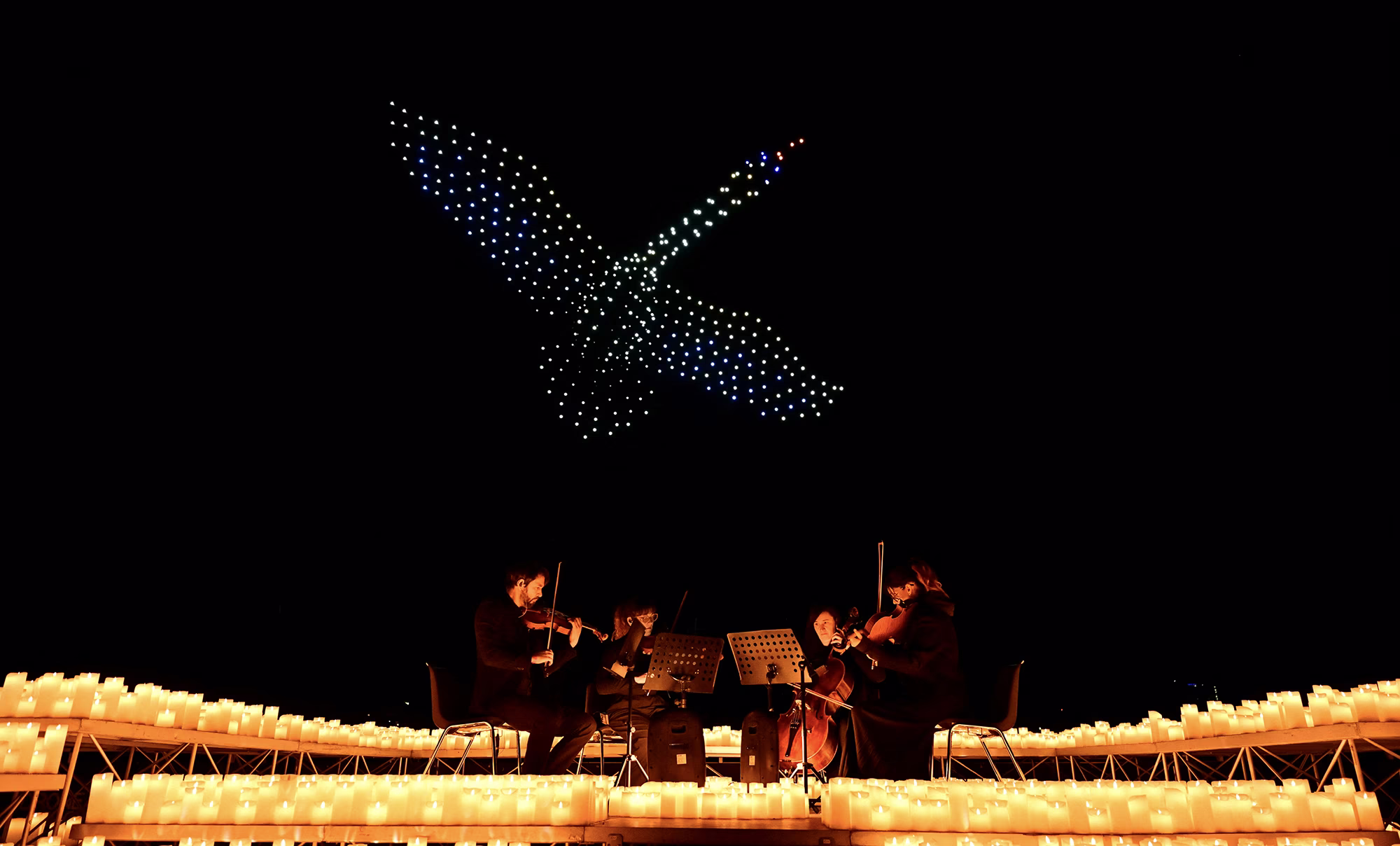 A string quartet performs as drones form the figure of a swan in the night sky above.