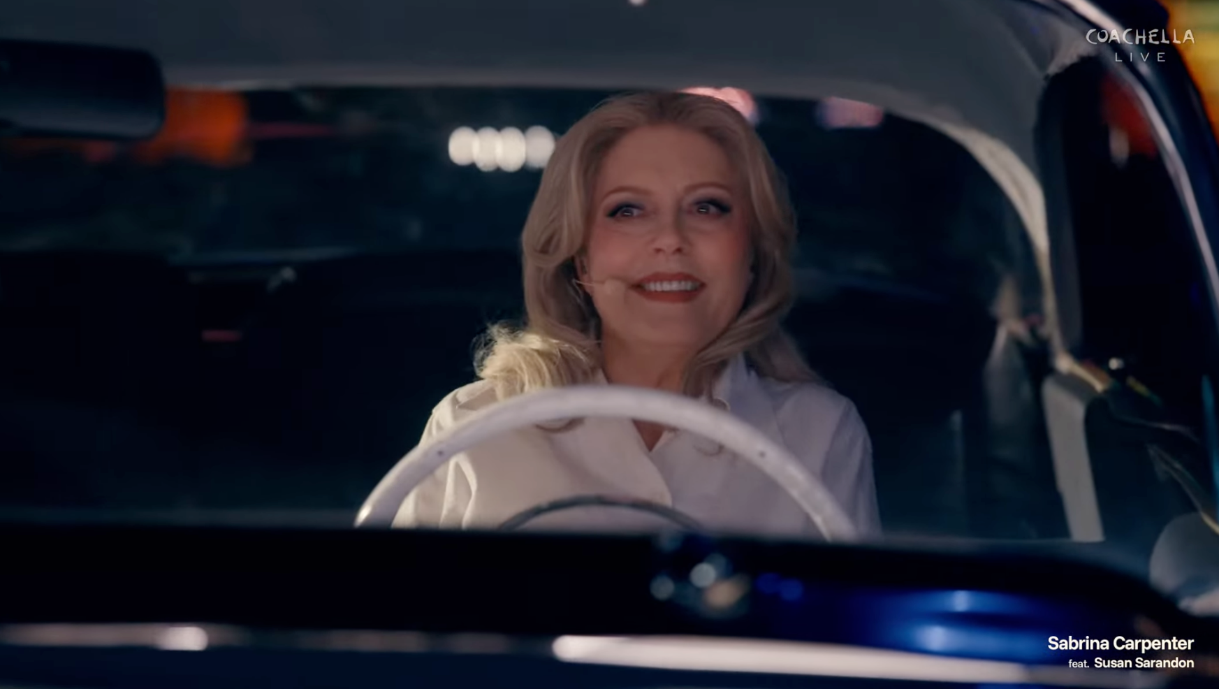 Susan Sarandon smiling in a white shirt behind the wheel of a car.