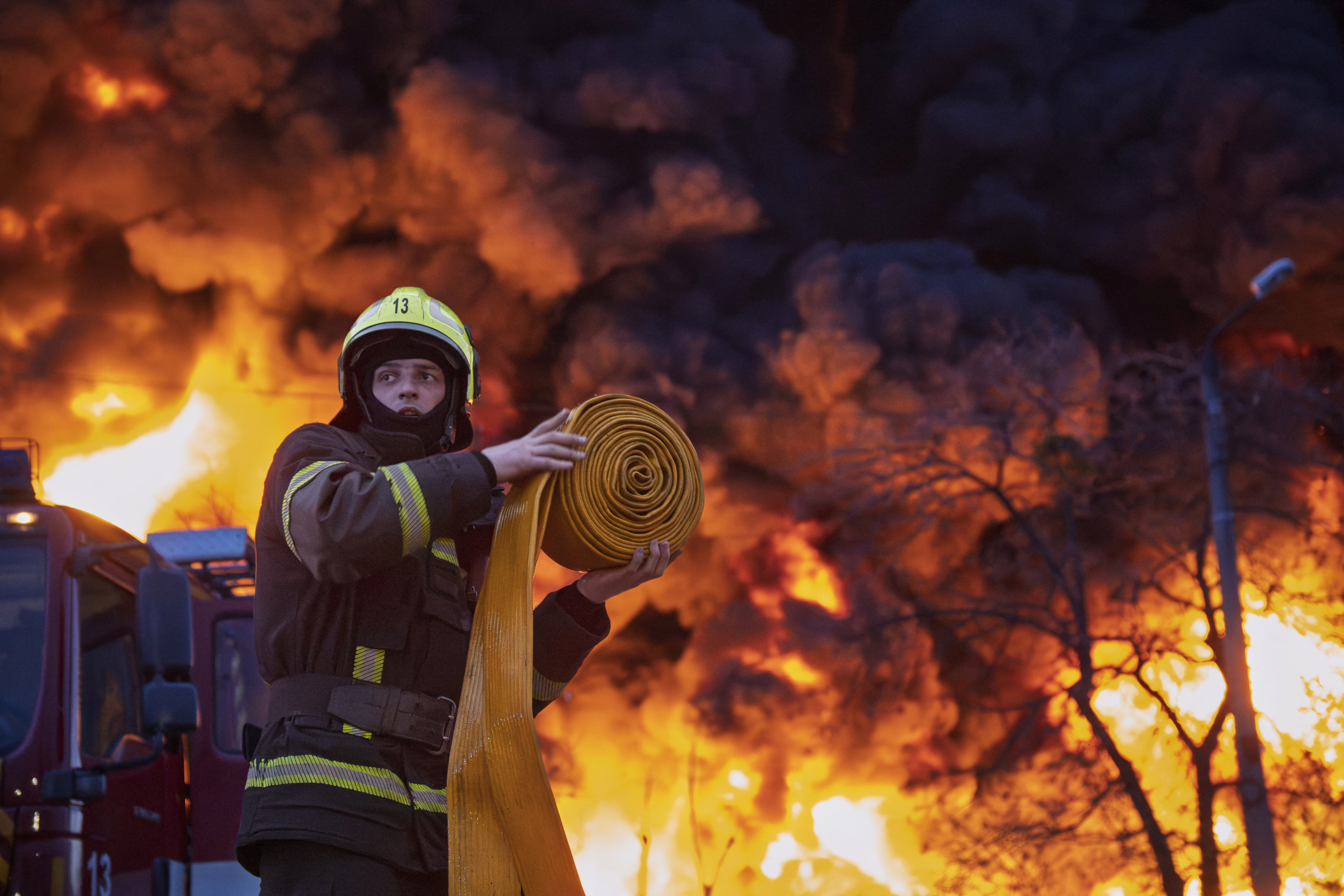 A firefighter unrolls a firehose, as smoke and flames rise up behind them.