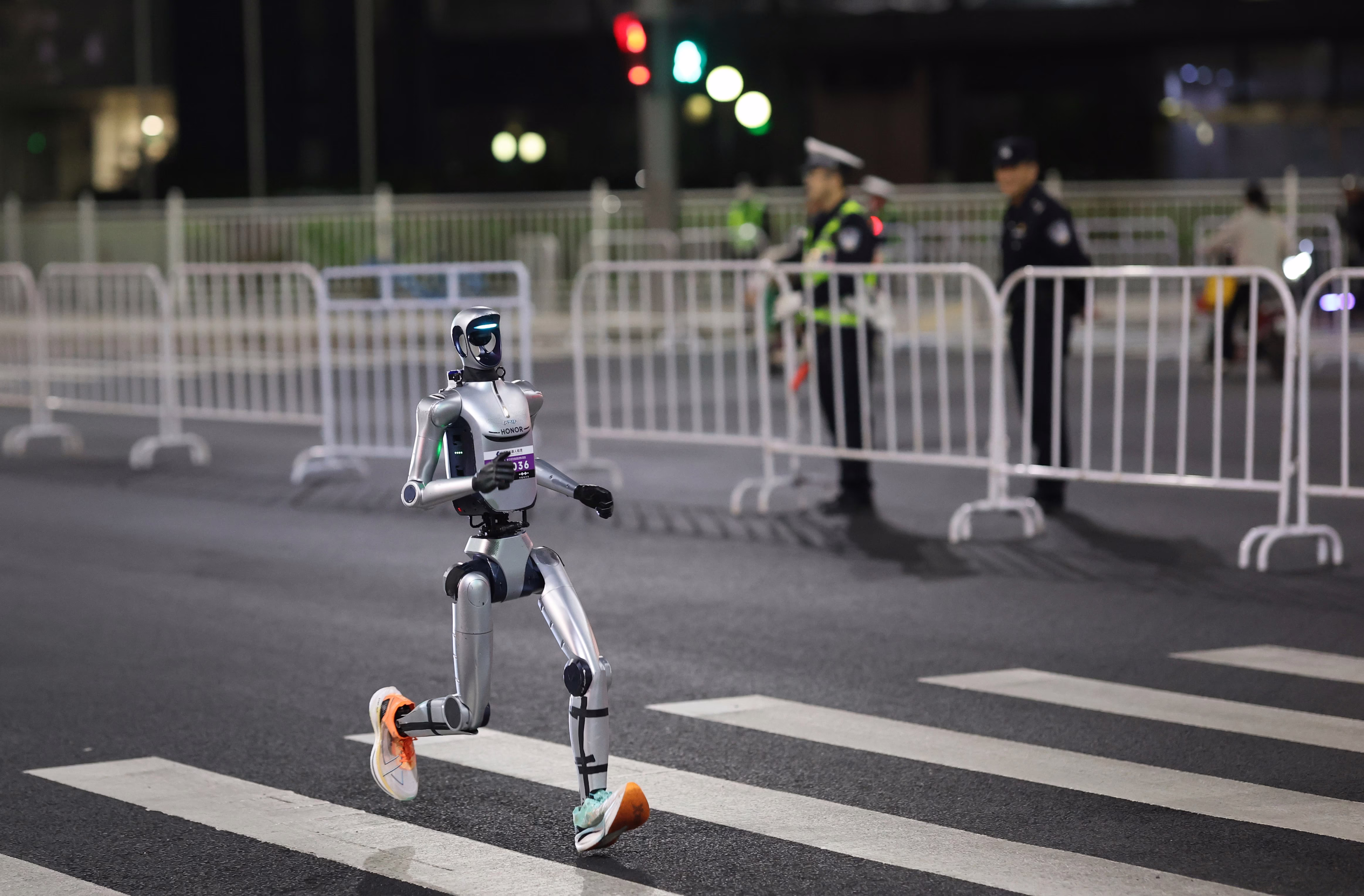 A humanoid robot wearing sneakers runs across a crosswalk, near several police officers standing beside barriers.