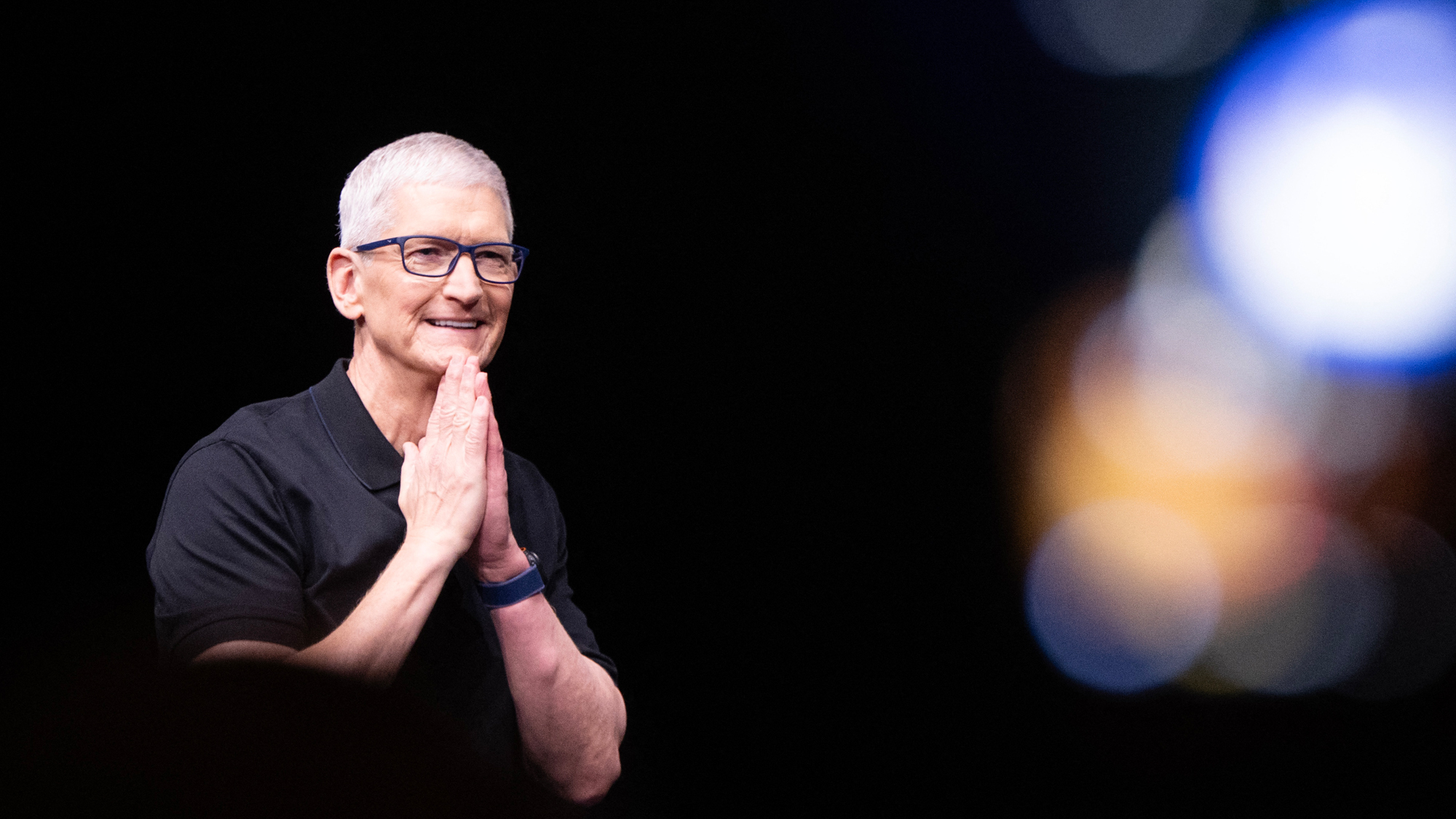 Tim Cook puts his hands together, touching his chin with them both. Blurry lights in the right part of the background