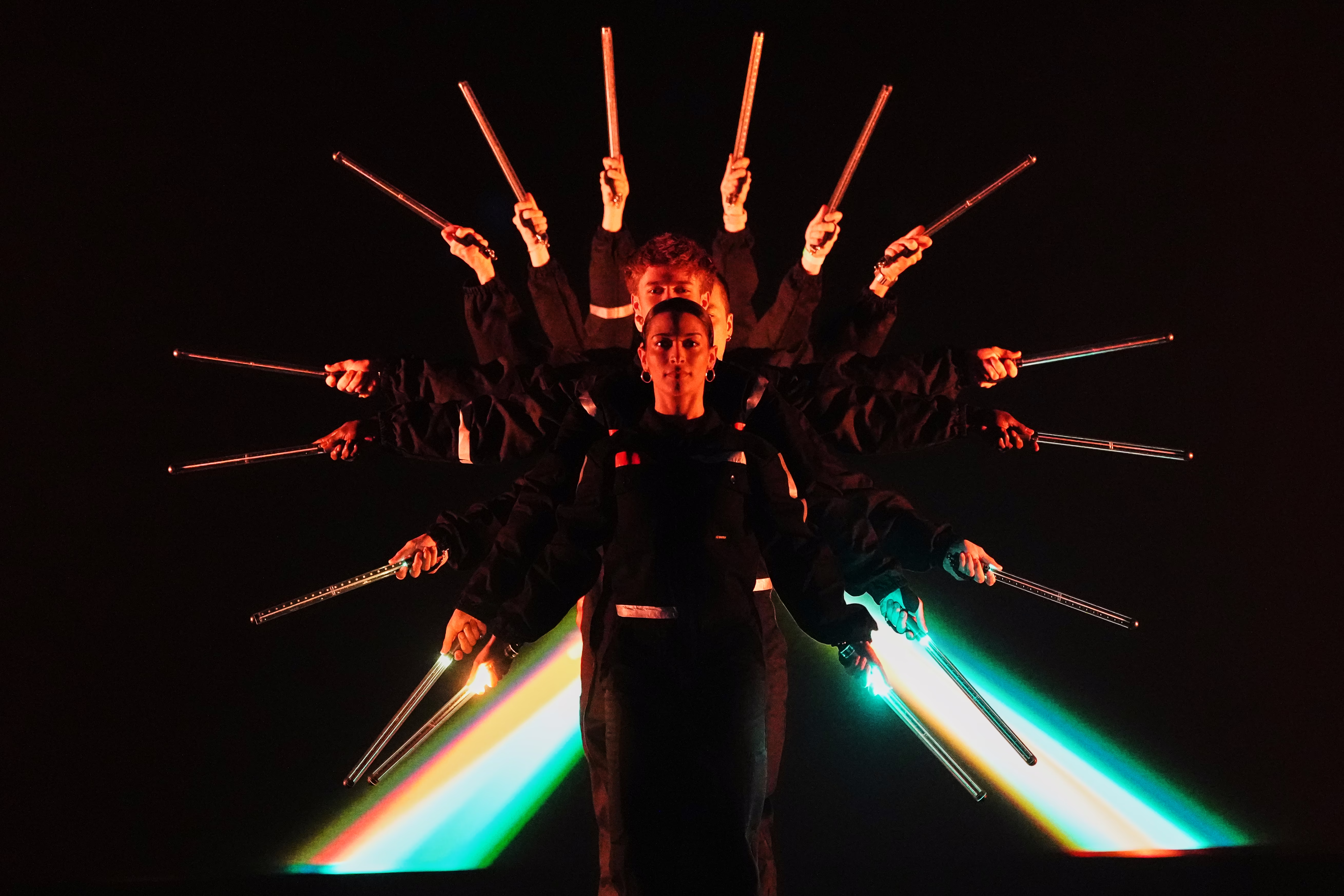 Eight performers pose onstage, facing forward, lined up closely front-to-back, each extending their arms at differing angles, holding wands out, making a starburst pattern.