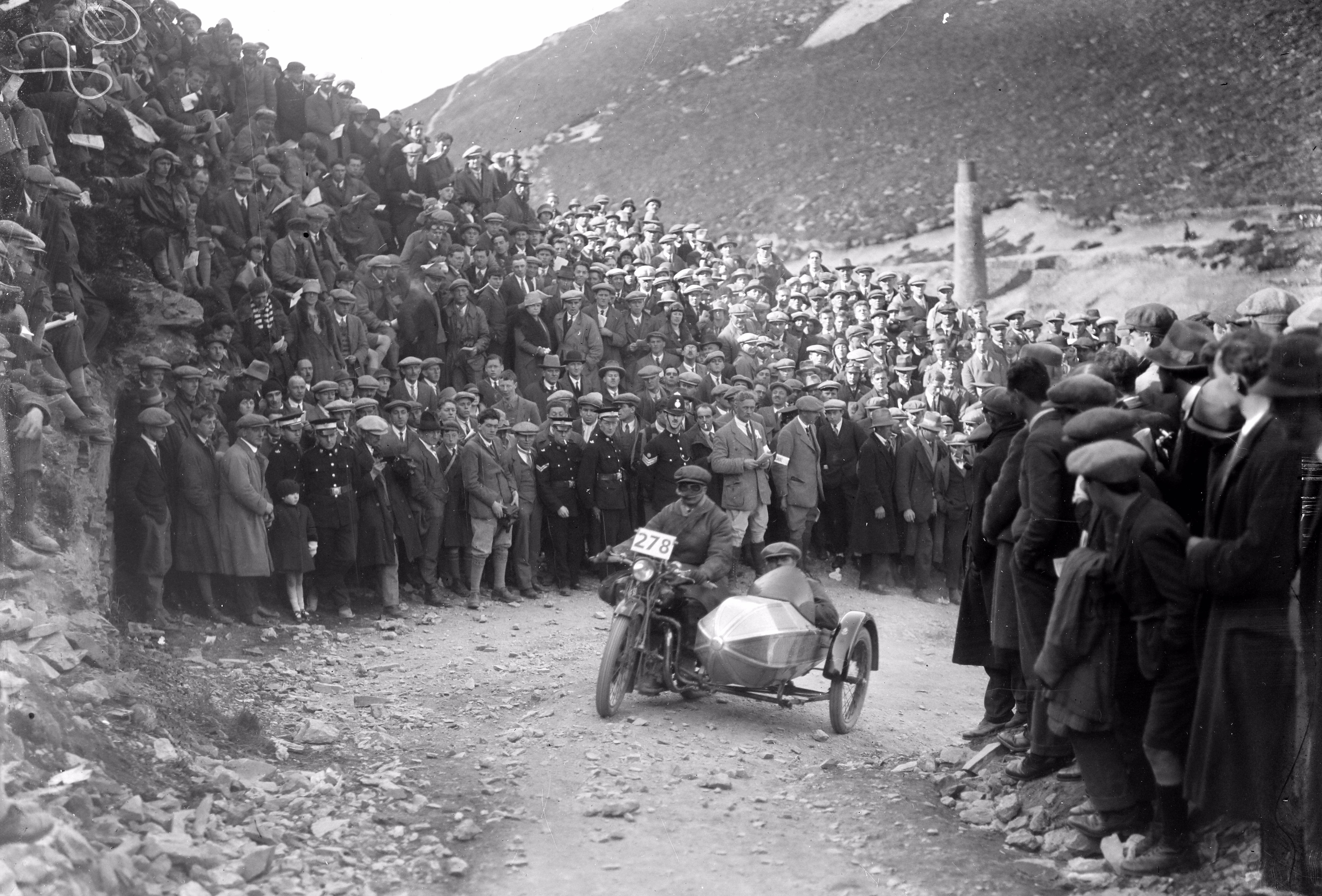 A large crowd of people stand on either side of a rough mountain road as a motorcycle with a sidecar passes by during a race in 1927.