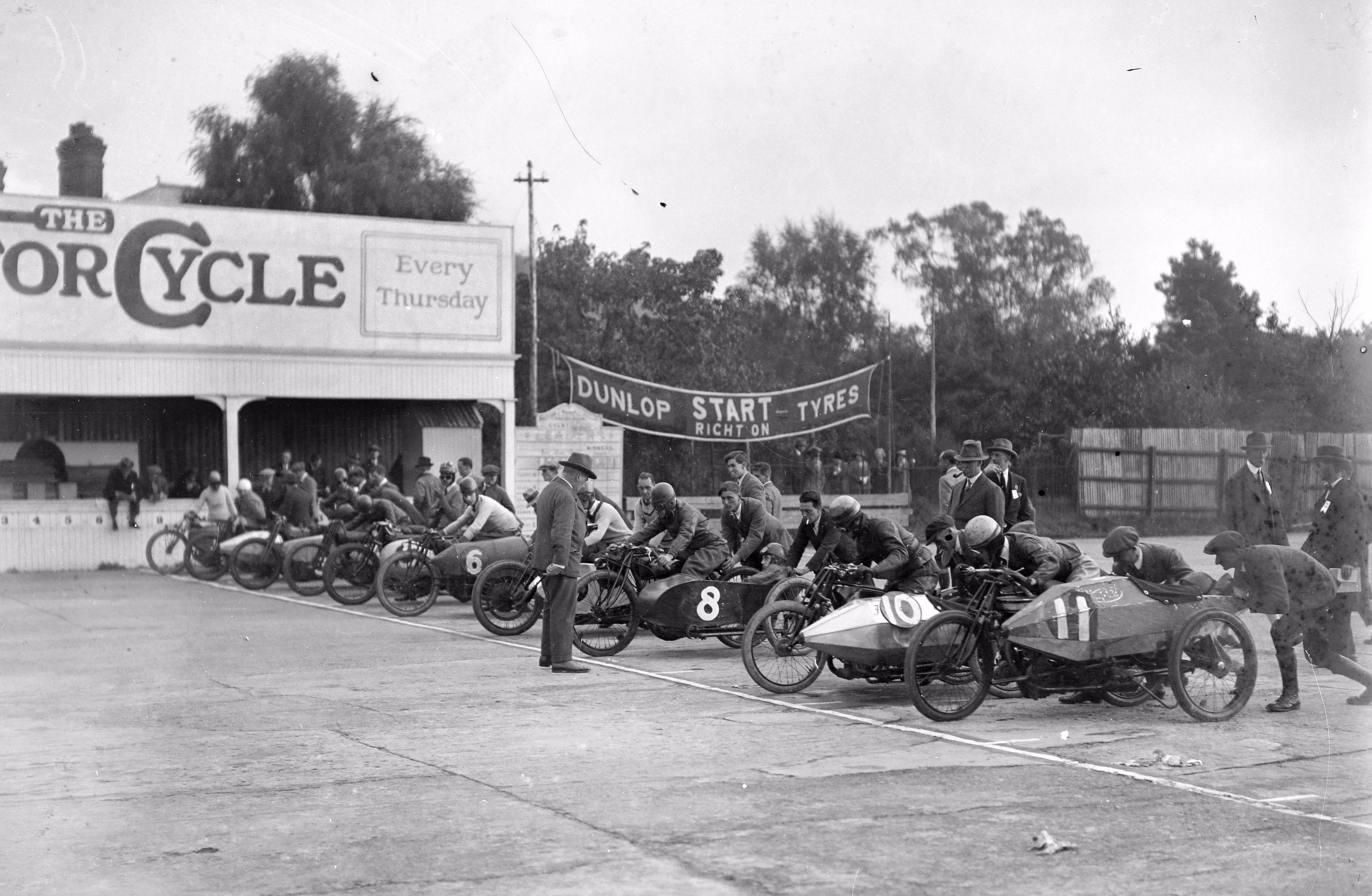 About a dozen motorcycles with sidecars are lined up at the start of a race in 1924.