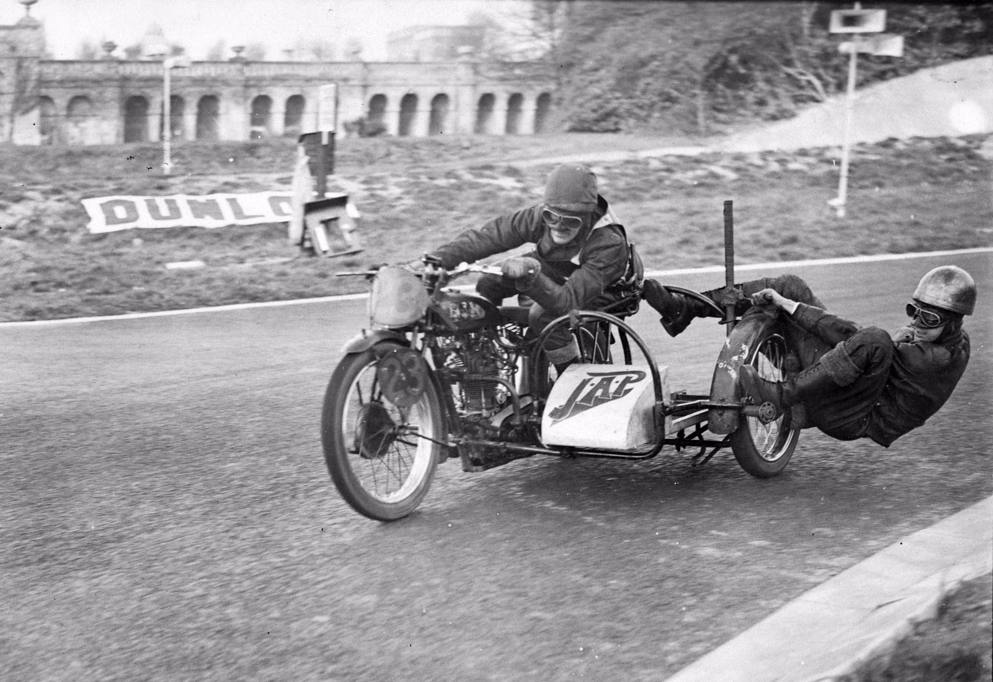 An early 20th-century black-and-white photograph of a motorcycle racer and his sidecar passenger leaning in a tight turn.