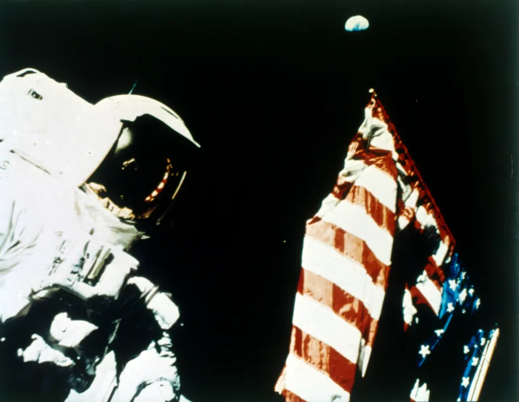 Astronaut Harrison Schmitt stands next to a US flag on the Moon, with Earth visible above the flag.