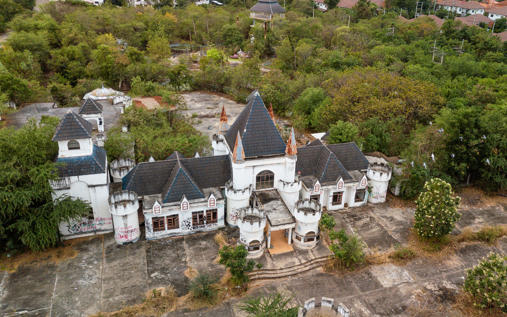 Aerial view of Castle Mall, an abandoned entertainment park in Thailand, showing a decaying, castle-like building surrounded by overgrown trees and concrete pavement.