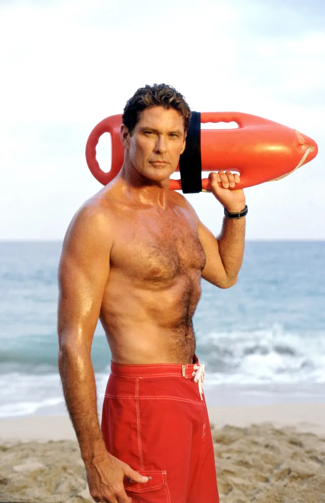 David Hasselhoff, bare-chested and in red swimming trunks, holds a red floatation device on his shoulder on a beach.