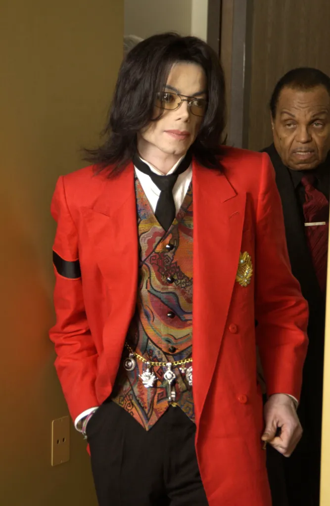 Michael Jackson, in a red blazer, patterned vest, and glasses, walks past the camera with Joe Jackson visible in the background.
