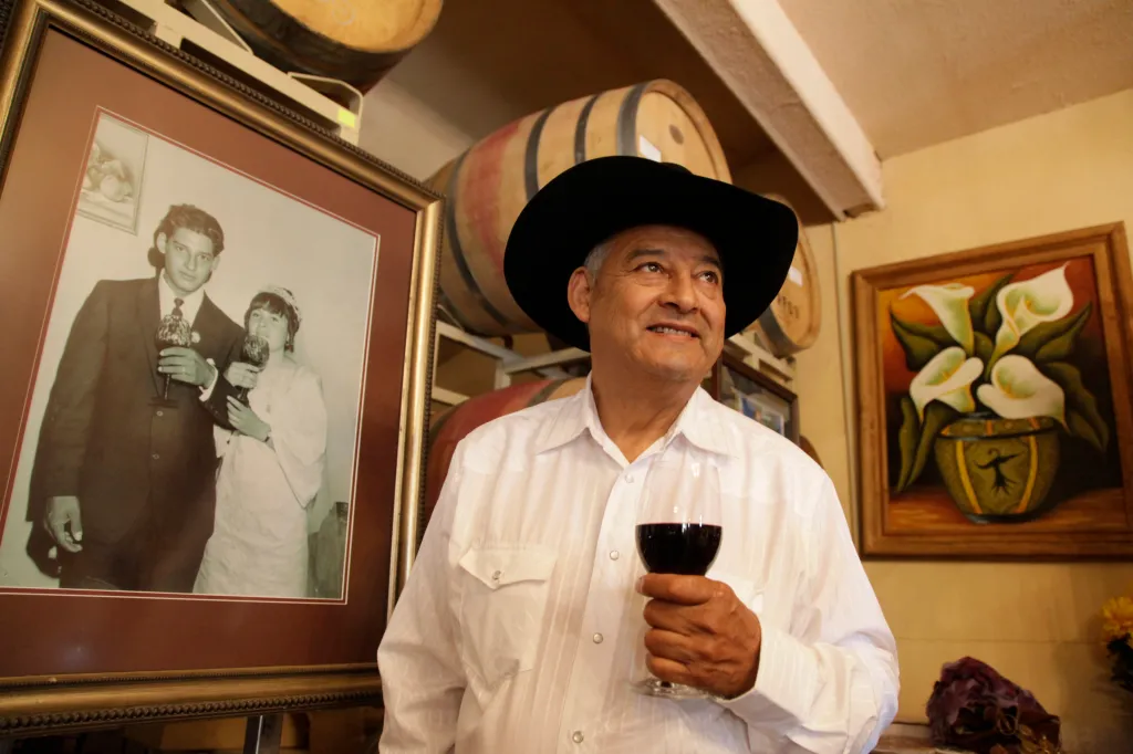 Reynaldo Robledo in his winery tasting room, holding a glass of wine, with a framed wedding picture of his younger self and his wife to his left.