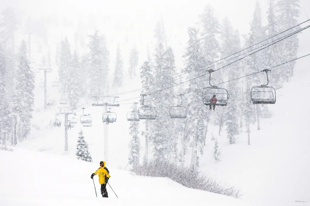 A skier in a yellow jacket stands on a snowy slope as chairlifts carry skiers in the background.