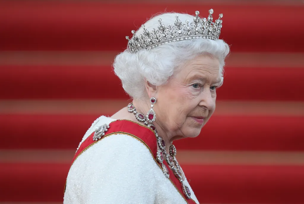 Queen Elizabeth II wearing a diamond tiara, necklace, and earrings with a red and white sash over a white gown.
