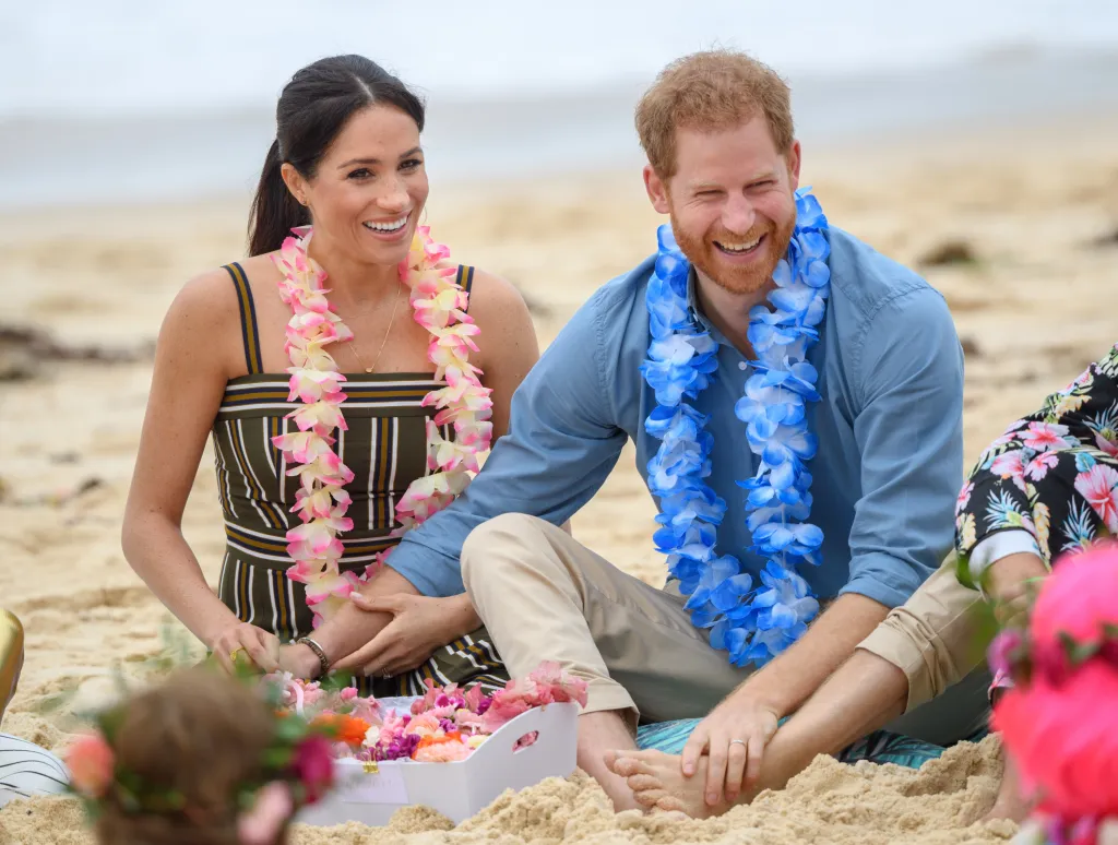 Prince Harry, in a blue shirt, blue floral lei and khaki pants, and Meghan Markle, in a brown and white striped dress, pink floral lei, wearing leis while sitting on Bondi Beach.