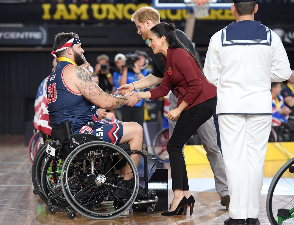 Meghan, Duchess of Sussex, in a dark red shirt and black pants, and Prince Harry, Duke of Sussex, in a blue shirt and grey pants, shaking hands with a wheelchair basketball player from Team USA at the Invictus Games.