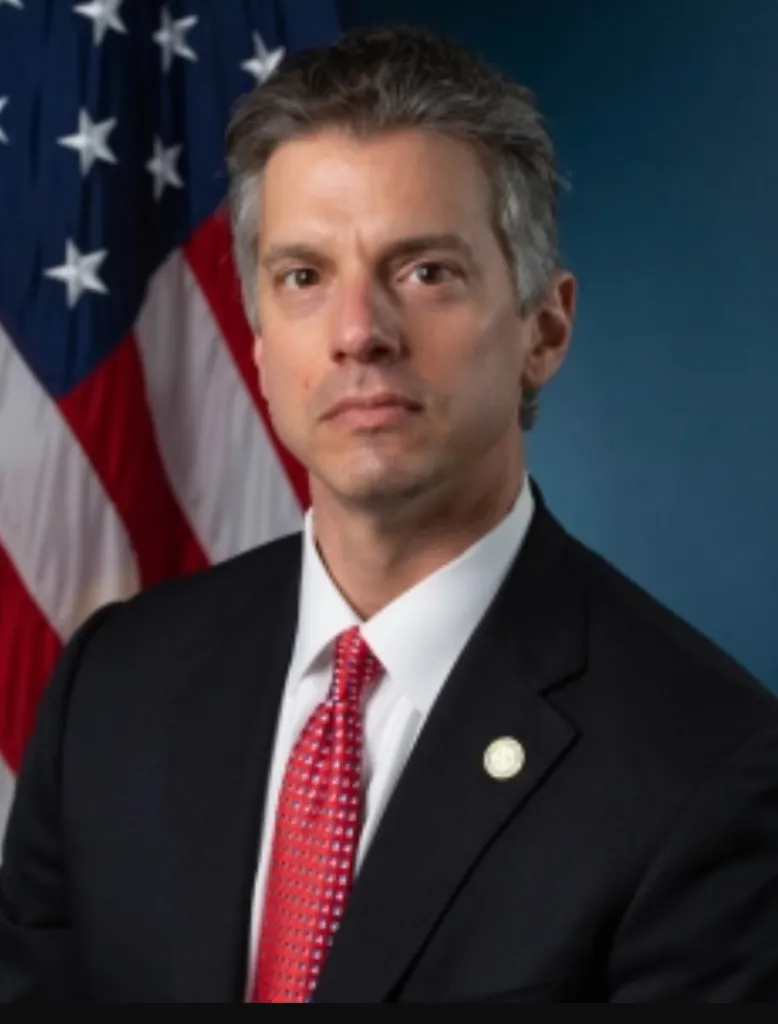 Adam Gordon, Assistant U.S. Attorney, wearing a suit and red tie in front of an American flag.
