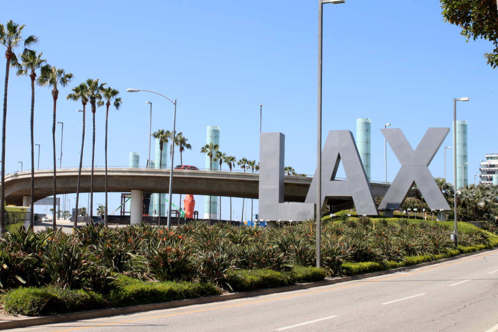 The iconic LAX sign, made of large gray letters, with palm trees and a road in the foreground and a bridge and terminal buildings in the background.