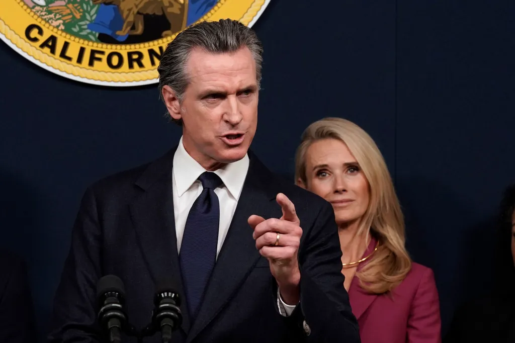 Gavin Newsom, wearing a suit and tie, speaking at a podium while Jennifer Siebel Newsom looks on behind him.