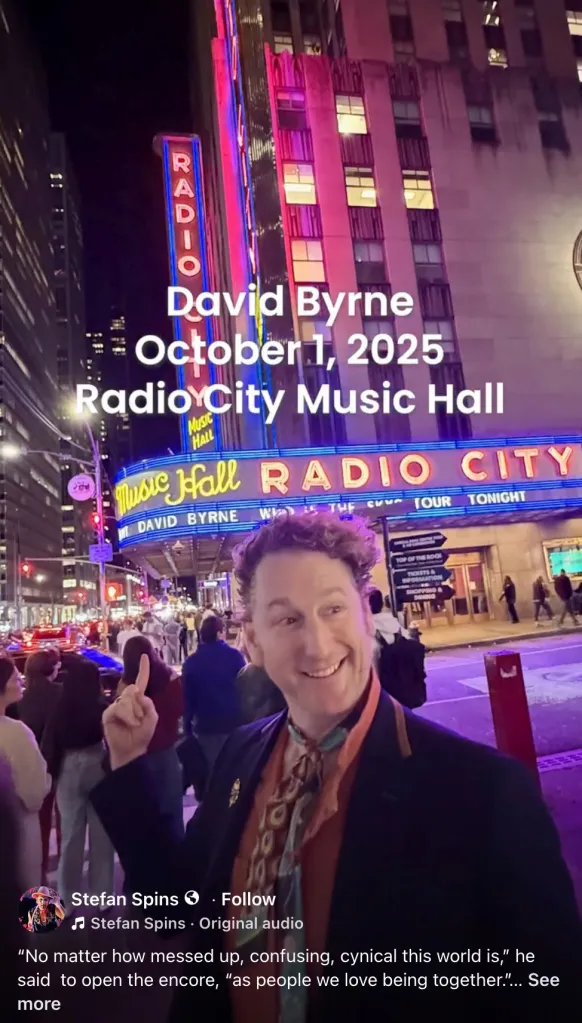 A man in a scarf points up at the bright neon sign of Radio City Music Hall in New York City, where text announces a 
