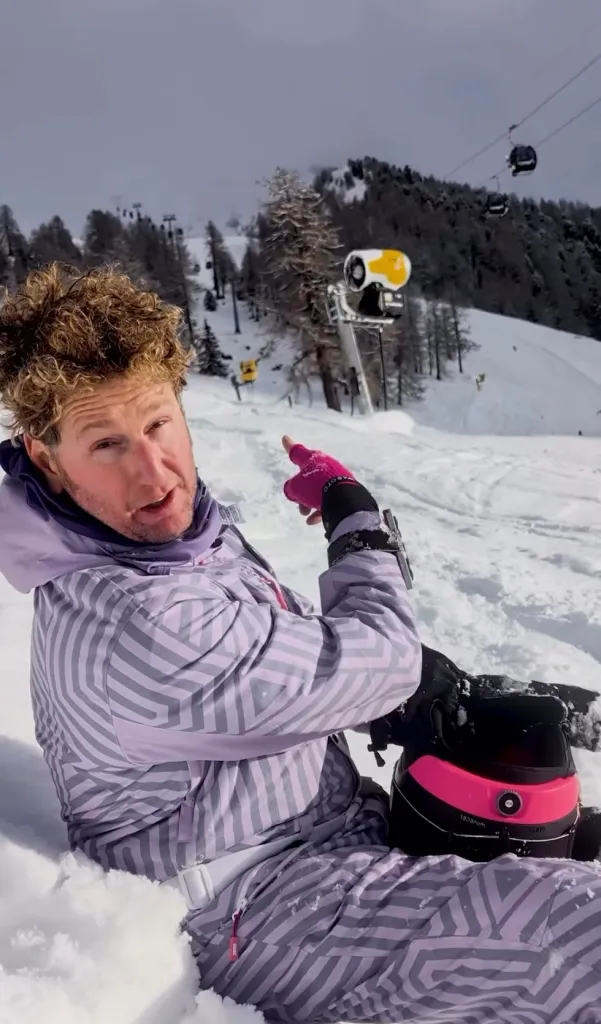 Stefan Pildes in snow, wearing a purple and gray patterned ski suit, pointing into the distance on a snowy mountain with trees and gondolas.