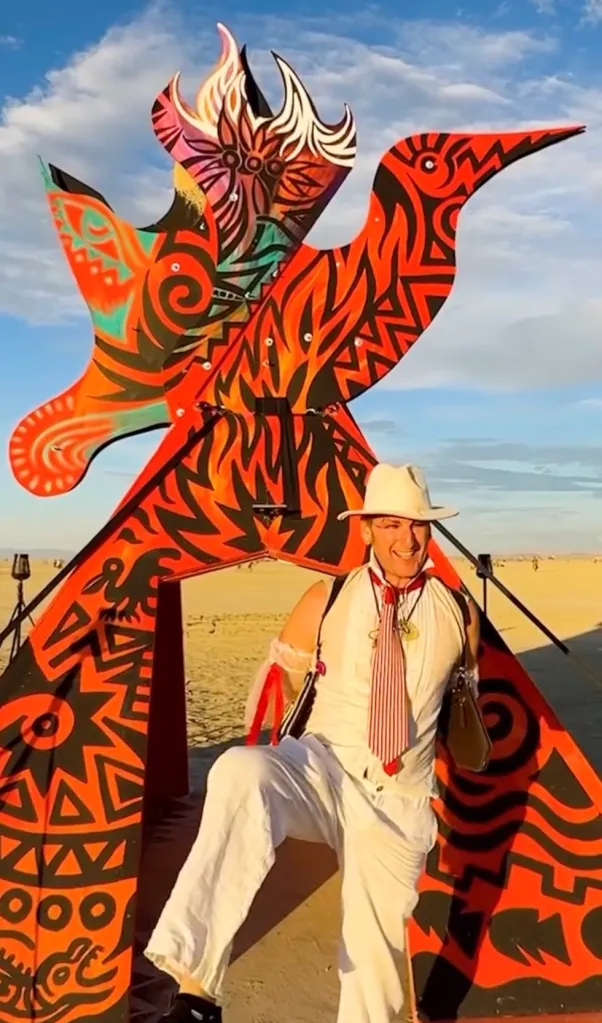 Stefan Pildes in white attire standing in front of a large, colorful bird sculpture in the desert at Burning Man.