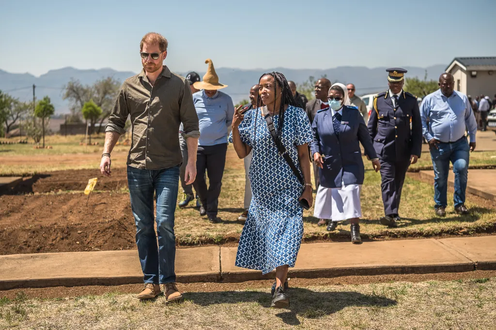 Prince Harry and Ntoli Moletsane, Sentebale Country Director in Lesotho, walk together, followed by other people, outdoors on a sunny day.