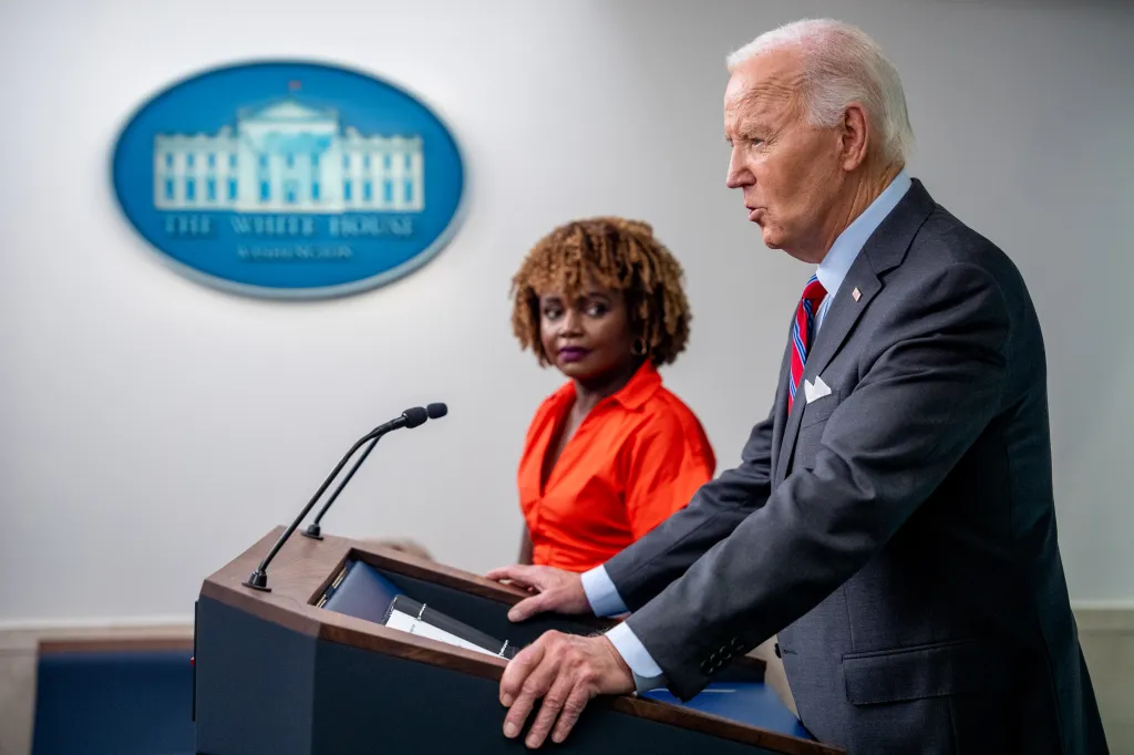 President Biden speaking at a podium with Press Secretary Karine Jean-Pierre in the background.