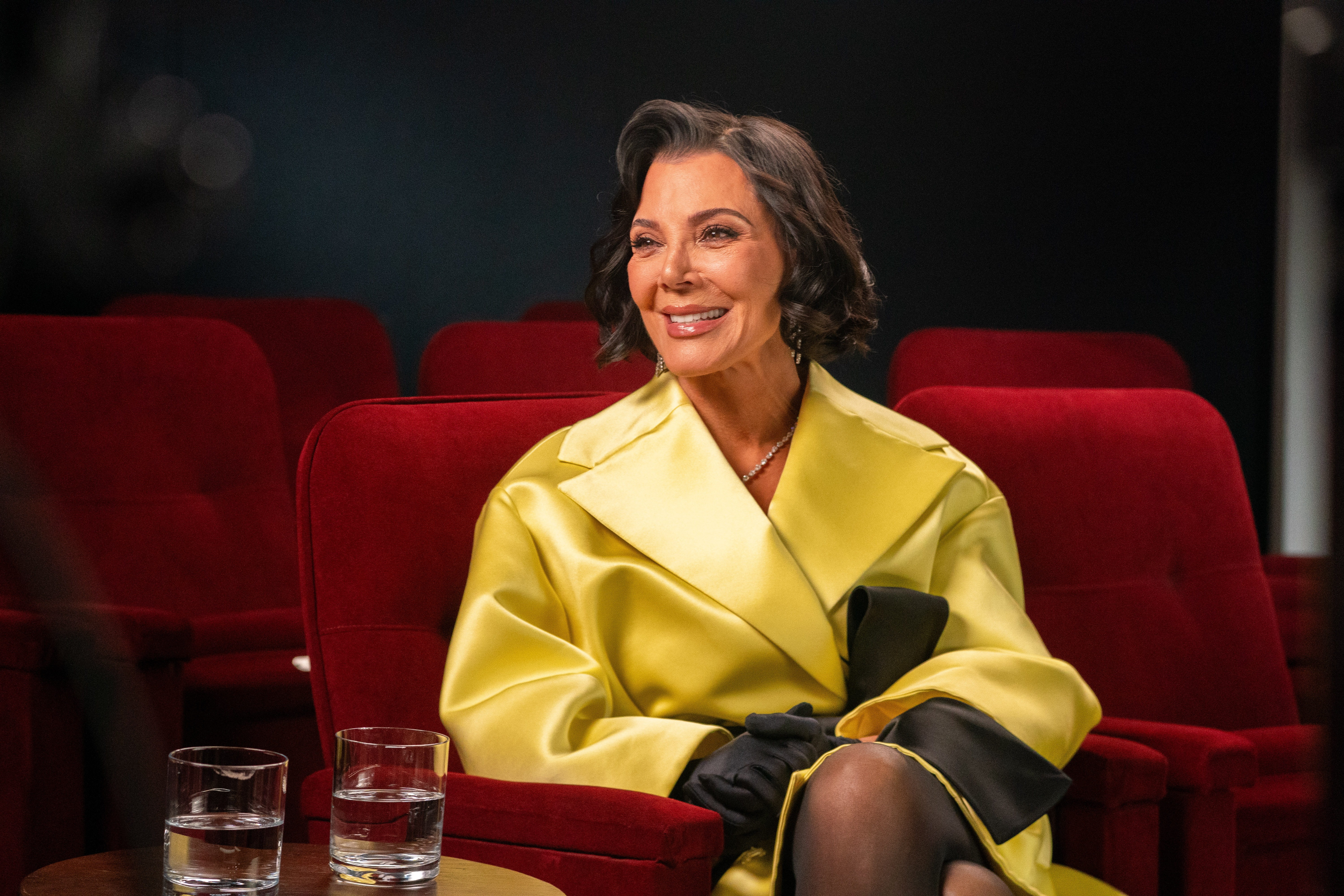 Kris Jenner sitting in a red velvet theater seat, wearing a bright yellow coat and black gloves.