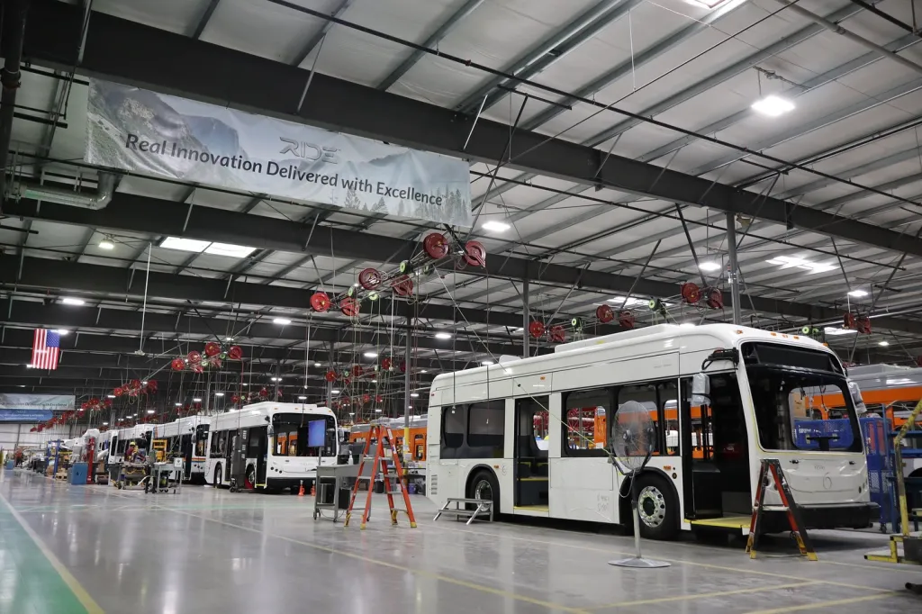 The RIDE bus factory in Lancaster, CA, with several white transit buses in various stages of assembly on the factory floor.