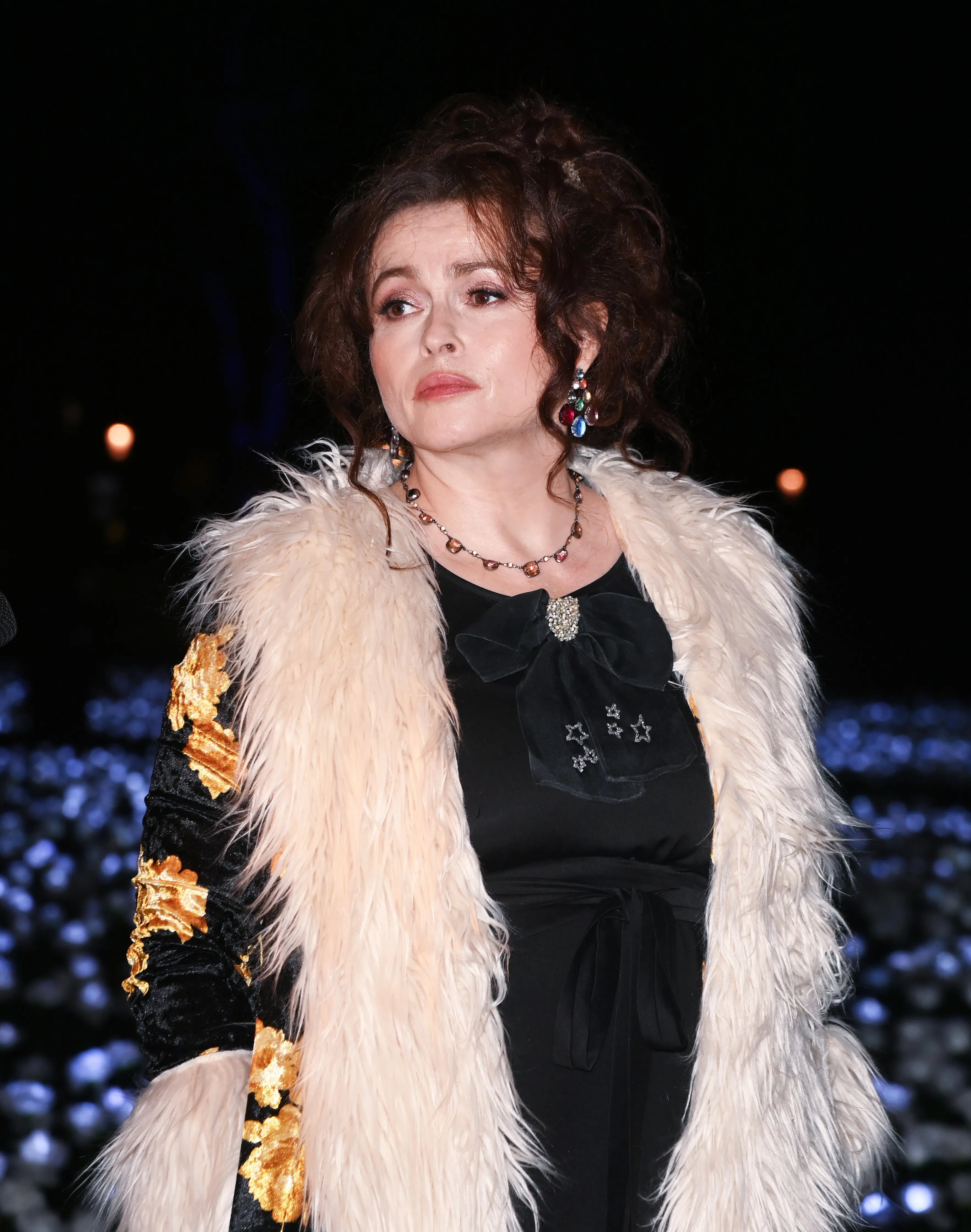 Helena Bonham Carter in a black dress with a bow, a light fur coat, and a gold-patterned sleeve, at the Ever After Garden event.