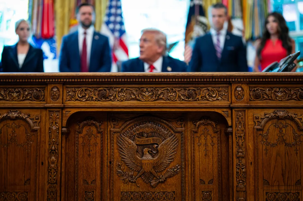 The Resolute desk with Donald Trump and others in the background.