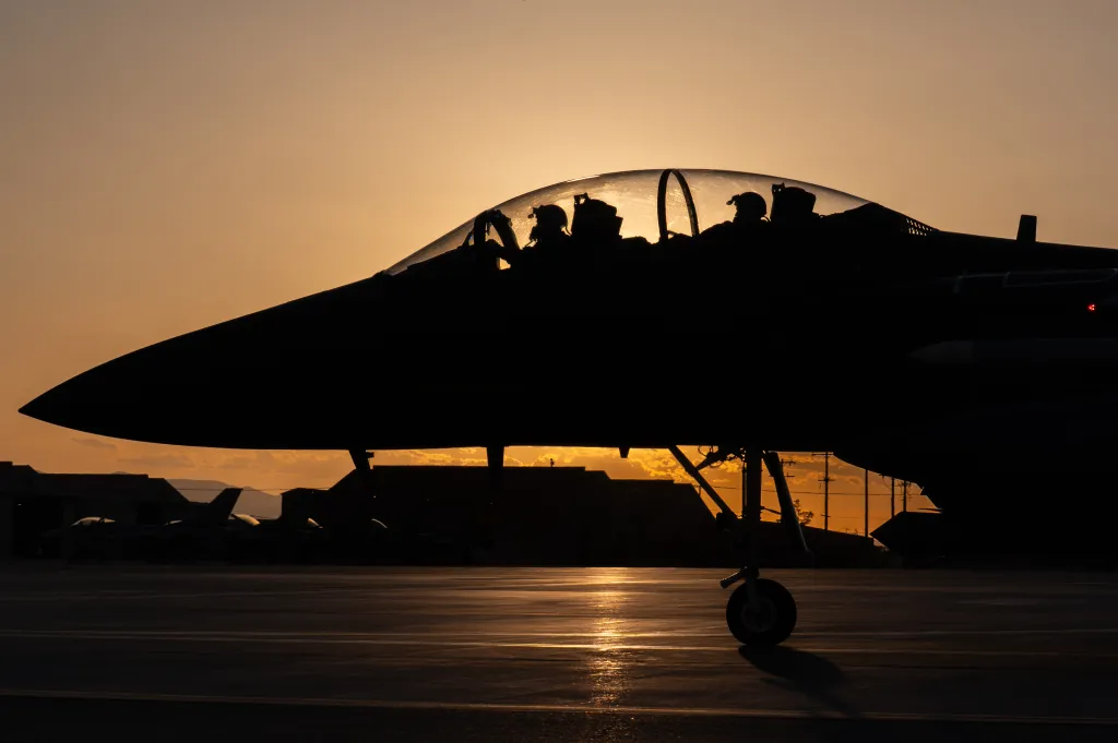 Silhouette of an F-15E Strike Eagle with two pilots in the cockpit, taxiing at Nellis Air Force Base at sunset.
