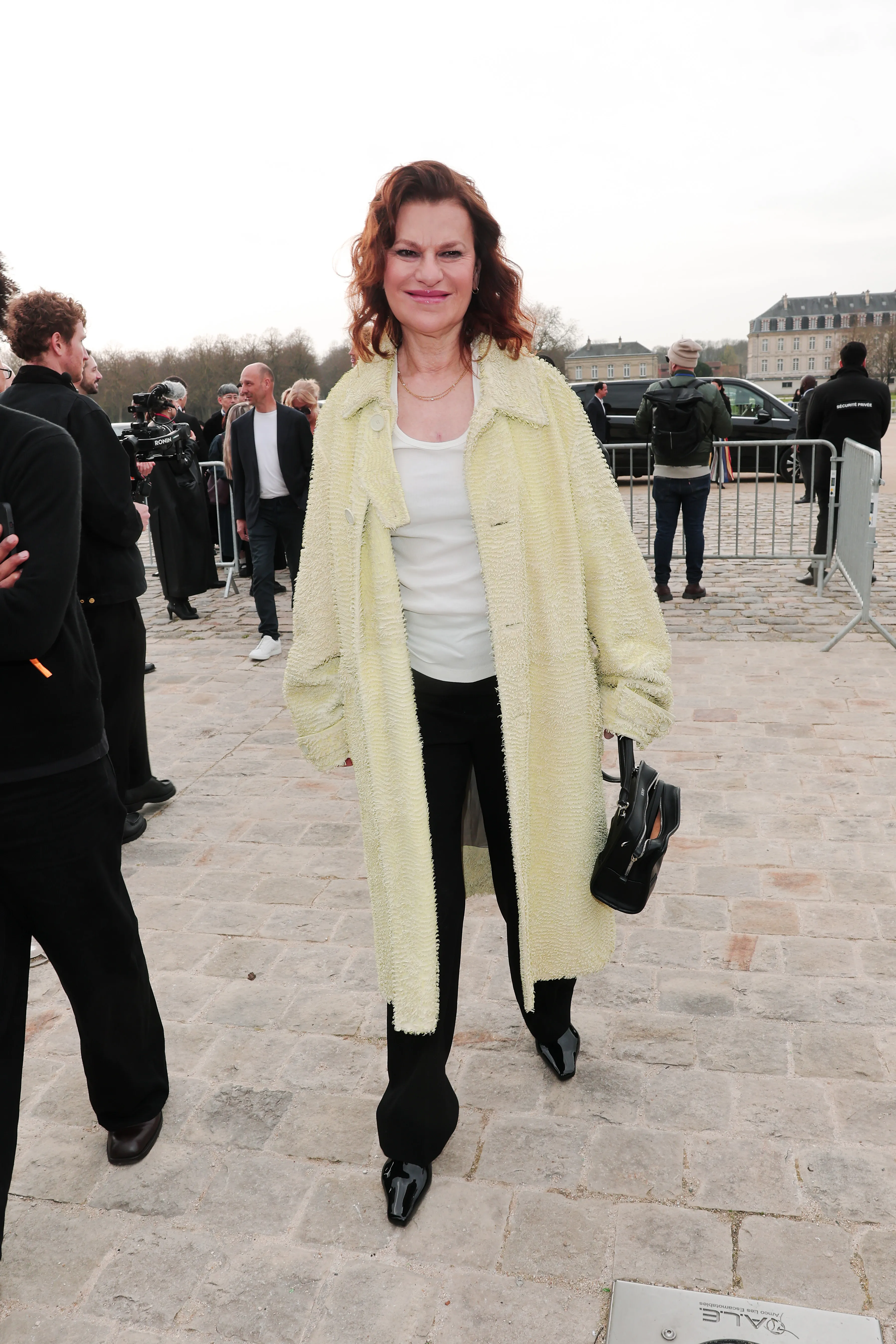 Sandra Bernhard wearing a yellow textured coat over a white top and black pants.