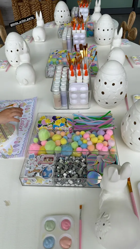Easter craft station with white ceramic bunnies and eggs, paints, brushes, pom-poms, stickers, and glitter.