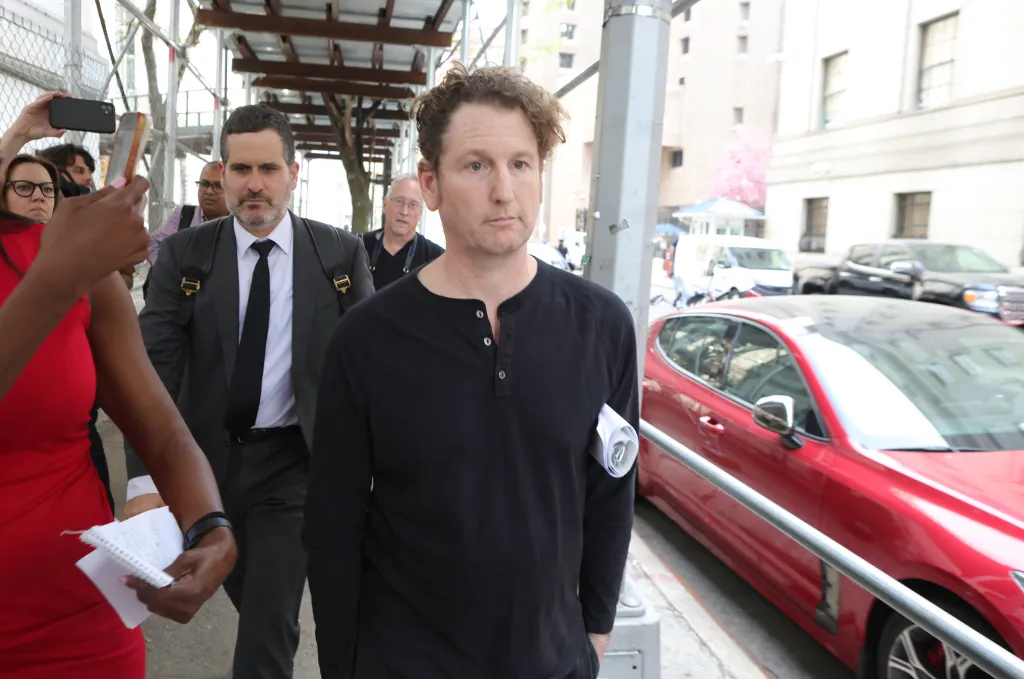Stefan Pildes, wearing a black shirt, walks on a city street with another man in a suit, while a crowd of people with phones record him.