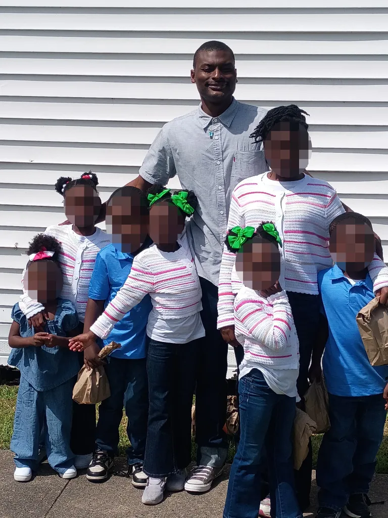 Shamar Elkins stands with his eight children, whose faces are blurred out, in front of a white building.