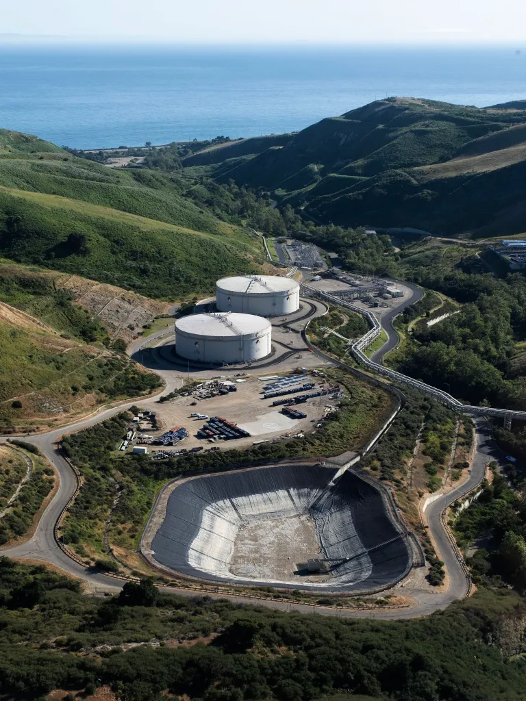 An aerial view of the Sable Offshore's Las Flores Canyon facility, with two large white storage tanks, an open lined pit, and roads winding through green hills to the ocean.
