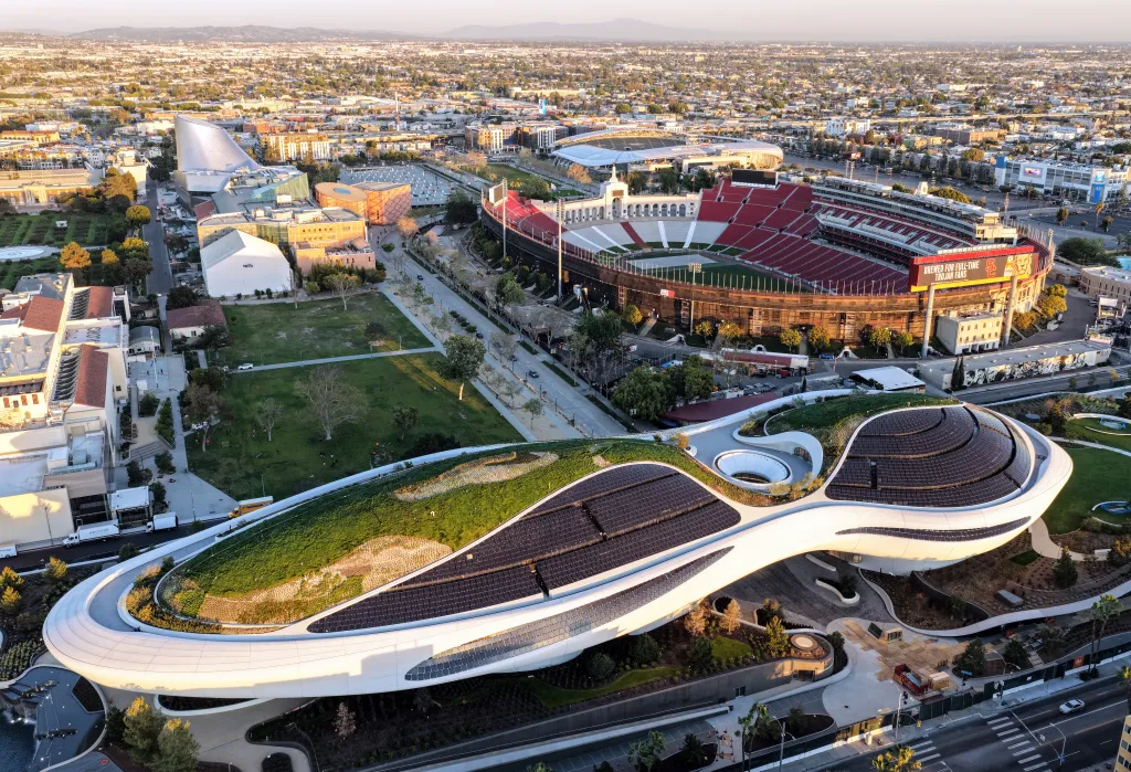 Aerial view of the Lucas Museum of Narrative Art with a green roof and solar panels in the foreground, with the Los Angeles Memorial Coliseum in the background.
