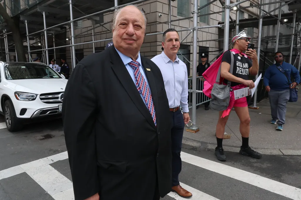 Billionaire John Catsimatidis smiling at the camera as he confronts a group of protesters outside his residence.