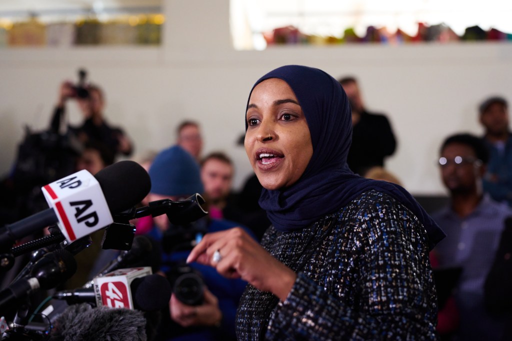Rep. Ilhan Omar speaking at a news conference, with microphones from various news outlets in front of her.