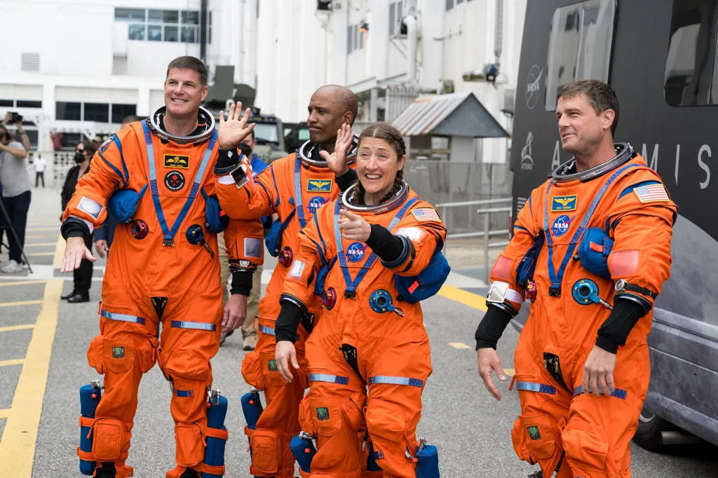 Four Artemis II crew members in orange spacesuits wave before boarding their Orion spacecraft.