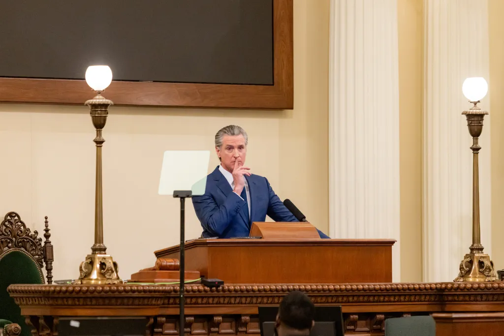 California Governor Gavin Newsom at a podium with his finger to his lips.