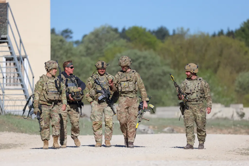Four soldiers in camouflage, carrying rifles and wearing helmets, walk on a dirt path during a military exercise.