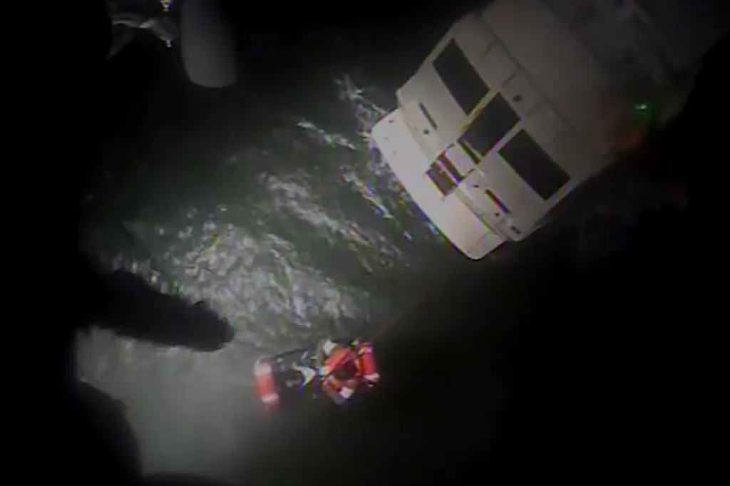 A Coast Guard rescue swimmer is hoisted from the water with a rescued person from a disabled motor yacht at night.