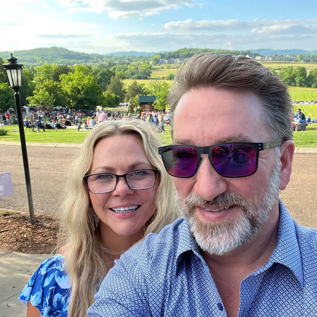 Chip and stepmom Amy McClure taking a selfie outdoors with a crowd of people and green hills in the background.