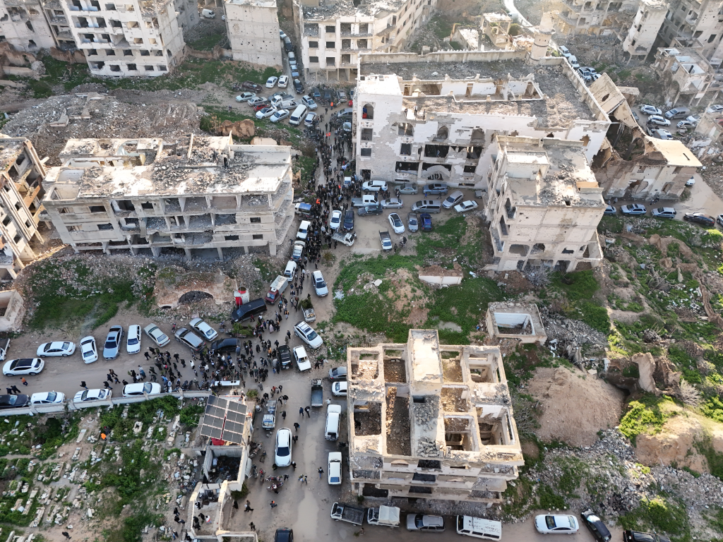 Aerial view of many people and cars in a war-torn city with severely damaged buildings.