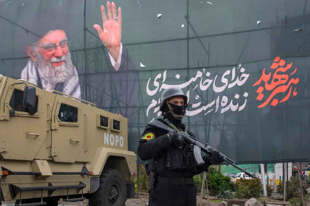 A soldier in black fatigues holds a rifle in front of a banner depicting Iran's Supreme Leader Ali Khamenei, with an armored vehicle nearby.