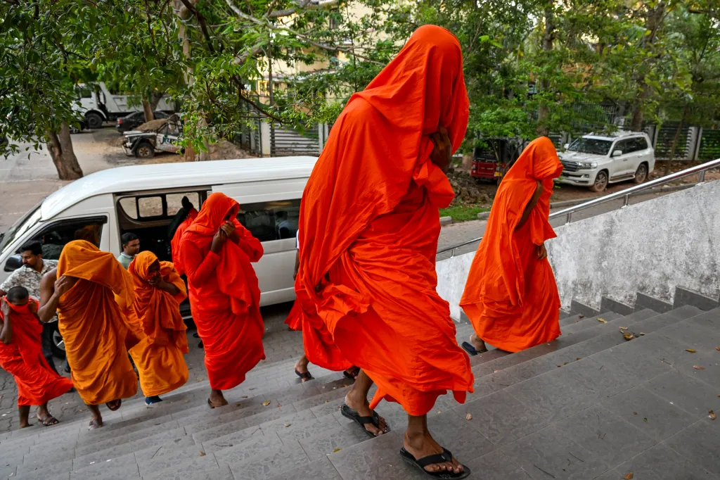 People dressed in orange robes walking up stairs with a white van and cars in the background.