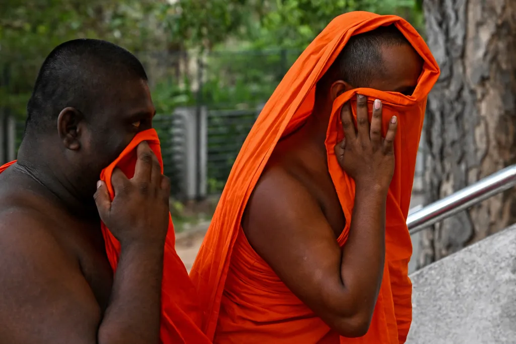 Two men in orange robes cover their faces, one partially with a cloth and the other fully with his robe.